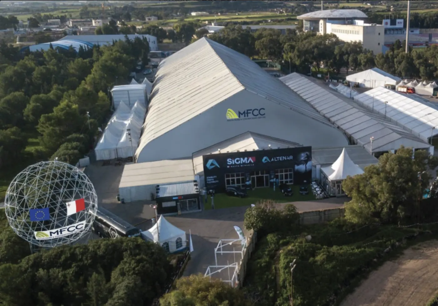 Aerial view of the Malta Fairs and Conventions Centre in Ta' Qali, its white pitched-roof hall ringed by marquees, with the geodesic entrance globe below